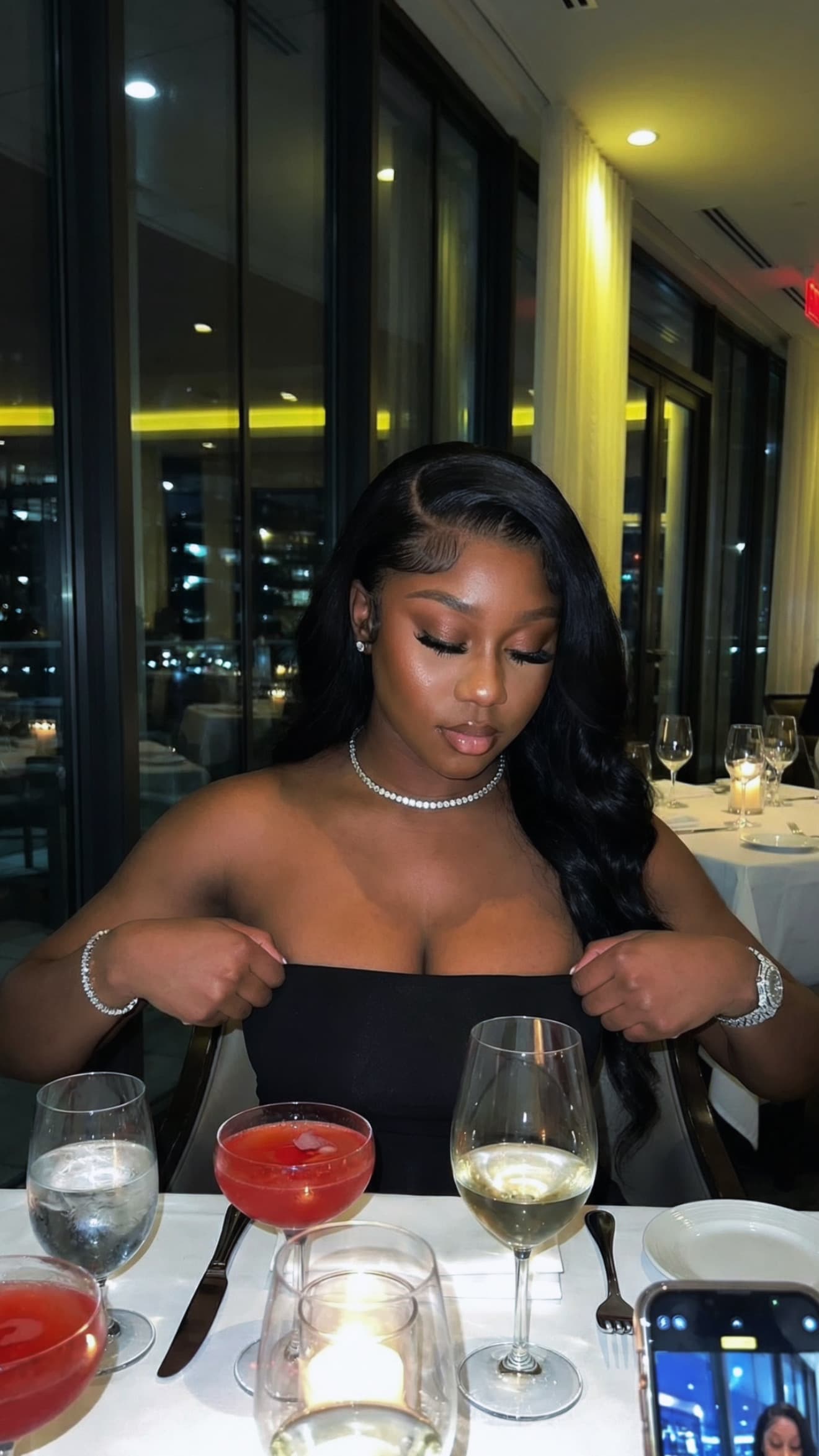 Elegant woman in a black strapless dress adjusts her top at a candlelit restaurant table.